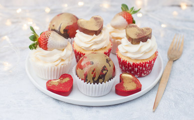 Cupcakes with cream cheese frosting decorated with fresh strawberries in white chocolate, chocolate red and golden balls and red chocolate hearts on the white plate. String lights on background.