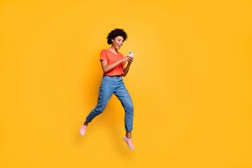 Fototapeta na wymiar Full length photo of positive cheerful funny curly brown hair girl use smartphone read post blogs in social network jump wear red t-shirt denim jeans isolated over yellow color background