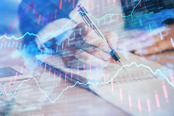 Obraz premium A woman hands writing information about stock market in notepad. Forex chart holograms in front. Concept of research. Multi exposure