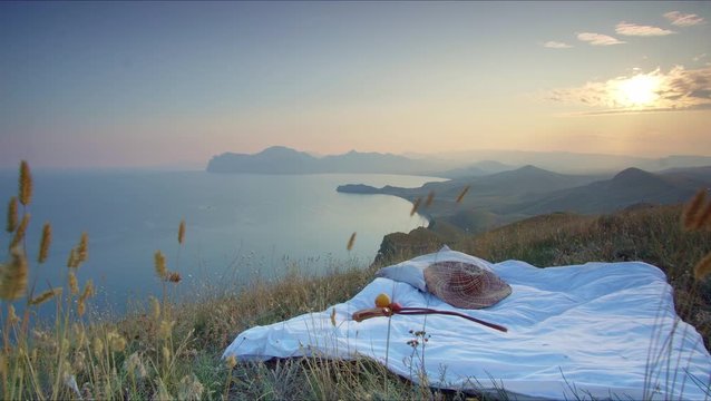beutiful shot on the sunset of blanket on the grass with hat and fruits on it, located on the top of the hill with amazing landscape and sea horisont