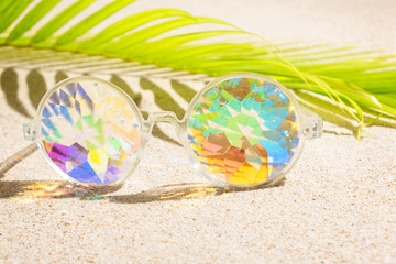 Funny kaleidoscope multicolour glasses on sand background with palm tree,  copy space, flat lay