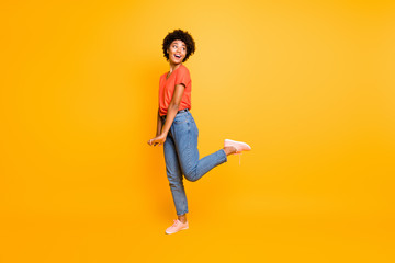 Full length body size photo of cheerful cute sweet girlfriend hesitating to look at what interested her wearing jeans denim orange t-shirt footwear isolated over yellow vivid color background