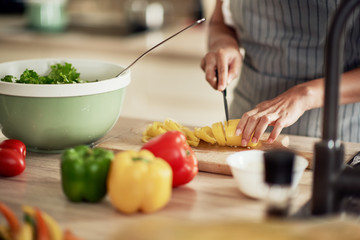 Close up of mixed race woman in apron cutting yellow pepper for dinner. Kitchen interior. On kitchen counter are peppers, zucchini and tomatoes.