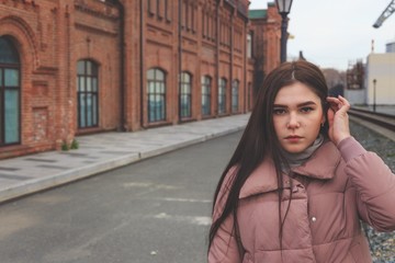 A young pretty pregnant girl in a pink warm down jacket walks near the old railway station. Late autumn, the cold is coming.