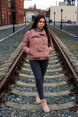 A young pretty pregnant girl in a pink warm down jacket walks near the old railway station. Late autumn, the cold is coming.