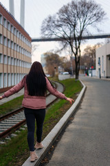 A young cute pregnant girl in a pink warm down jacket is trying to walk on a high curb. Late autumn, the cold is coming.