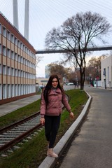 A young cute pregnant girl in a pink warm down jacket is trying to walk on a high curb. Late autumn, the cold is coming.