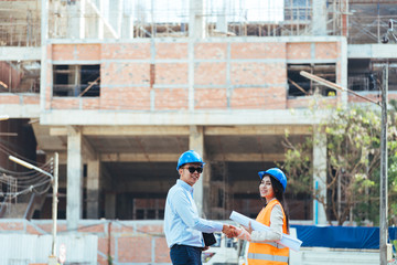 Asian man civil engineer and woman architect wearing blue safety helmet meeting at contruction site.