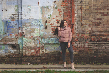 Young cute pregnant girl in pink warm down jacket looks away.  Her hand is touching her hair.  Late autumn, the cold is coming. Old brick wall on the background. toned photo.