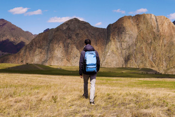 tourist in a cap and jacket with a backpack goes on the grass in the direction of mountains....