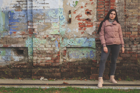 Young cute pregnant girl in pink warm down jacket looks away.  Late autumn, the cold is coming. Old brick wall on the background. toned photo.
