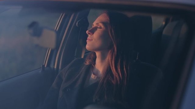 Cute female sit in the car and rest close her eyes in the evening at sunset. Beautiful woman sleeping in car in the evening sun. Shooting through windshield.