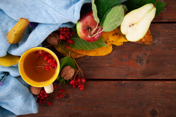 Cropped photo of a white cup with tea and a violet sweater strewn with orange maple leaves on a wooden table. The concept of comfort, coziness and hygiene. Autumn mood, fall season. Selective focus,