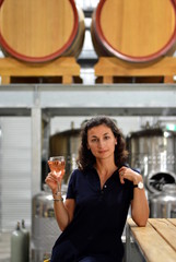 A young beautiful smiling woman sitting on a high chair and holding a glass of rose wine in a winery with wine barrels on a background, Hunter Valley, Australia
