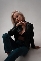 Fototapeta premium Hot trendy fashion woman model in leather jacket, jeans and underwear posing on white background in room. Portrait of sexy stylish hot blonde girl in lingerie with beauty face and body.
