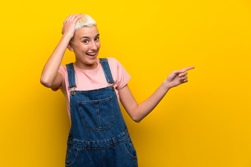 Teenager girl with overalls on yellow background surprised and pointing finger to the side