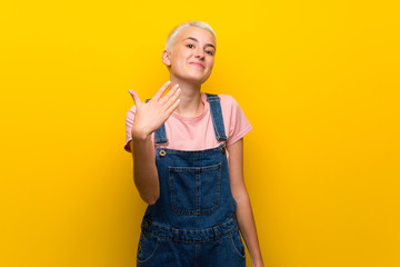 Teenager girl with overalls on yellow background inviting to come with hand. Happy that you came