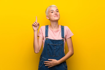 Teenager girl with overalls on yellow background showing and lifting a finger in sign of the best
