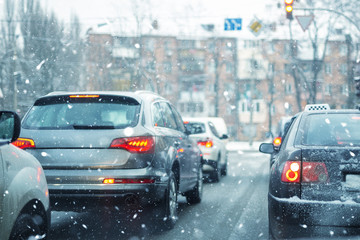 Many cars standing in row on traffic lights during snowfall in cold winter morning. Traffic jam on city street at snowstorm weather