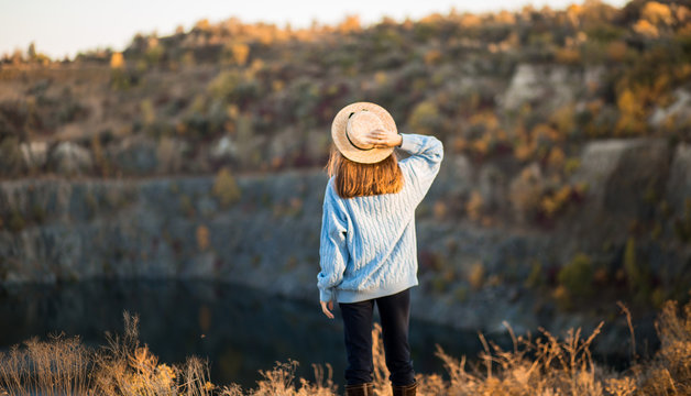 Autumn girl in blue sweater and hat standing backwards and admire nature lake view. Autumn forest colors with girl back view. Autumn portrait.