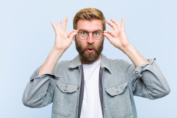 young blonde adult man feeling shocked, amazed and surprised, holding glasses with astonished,...