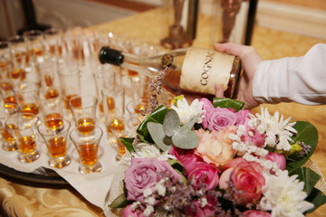 The bartender pours old cognac into a small shot glass. An Old bottle of cognac and Lots of small shot glasses on the table. Flowers Wedding table decoration.
