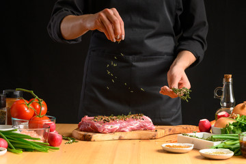 Pork steak and green herbs, rosemary and thyme from the chef in the background. Horizontal photo. With vegetables, tomatoes, hot peppers, onions and a chapel. With black background