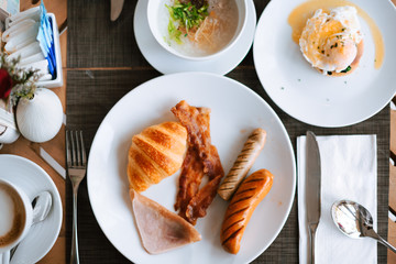 Top view of simple breakfast including ham, sausage, bacon, croissant, egg benedict, boil rice and coffee.