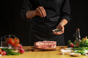 Chef cooks pork ribs. sprinkling with pepper on a background with vegetables. Freezing in motion. Recipe book, cooking, restaurant business