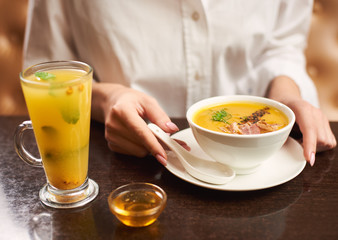 Woman in white blouse ordering tasty and delicious meal at restaurant. Female hands touching white bowl with orange cream soup. Near first dish on brown table standing tea with mint and honey.