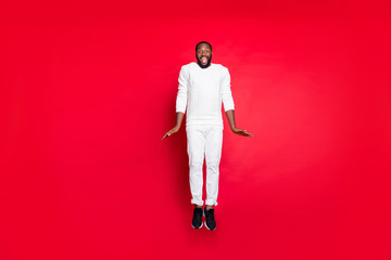 Full size photo of excited dark skin handsome guy jumping high rejoicing having best x-mas party time wear white sweater trousers isolated red background