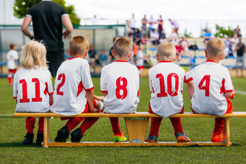 Sports team on the wooden bench during the youth tournament on a sunny summer day. Parents as...