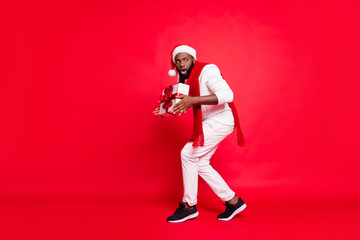 Fototapeta na wymiar Full body photo of dark skin man making newyear surprise for family going tiptoe carry giftbox under x-mas tree wear sweater trousers santa hat isolated red background