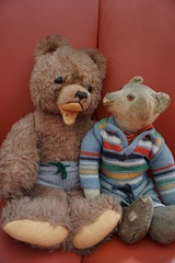 two old cute teddy bears are sitting on a red couch