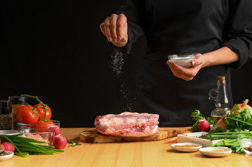 Chef cooks pork ribs. sprinkling with salt on a background with vegetables. Freezing in motion. Recipe book, cooking, restaurant business