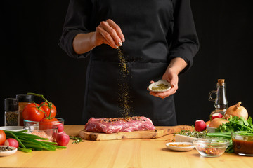 Pork steak and green herbs from the chef in the background. Horizontal photo. With vegetables, tomatoes, hot peppers, onions and a chapel. With black background