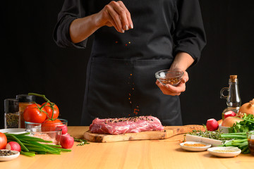 Pork steak and seasoning from the chef in the background. Horizontal photo. With vegetables, tomatoes, hot peppers, onions and a chapel. With black background