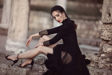 A beautiful girl in a long black dress with her legs open, poses at sunset amid antique columns, and ruins.