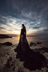 Fototapeta na wymiar A beautiful girl in a long black dress poses at sunset amid the sea. Beautiful sky.