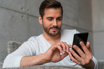 Blurry friendly young man in casual clothes using smartphone sitting in armchair in loft office or at home. Focus on hands. Concept of social media and web surfing.