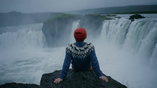 Man in red beanie and blue wool ornament sweater sits on edge of epic rock or cliff overlooking epic waterfall in iceland or norway. Magical and inspirational wanderlust concept
