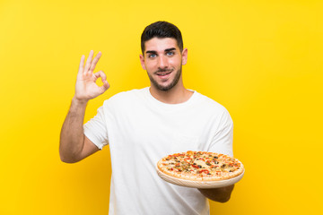 Young handsome man holding a pizza over isolated yellow wall showing ok sign with fingers