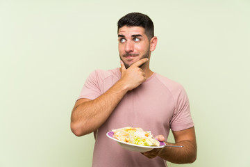 Young handsome man with salad over isolated green wall thinking an idea