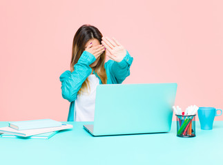 young pretty woman working with a laptop covering face with hand and putting other hand up front to...