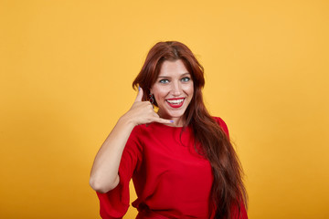 Gorgeous girl smiling, holding right hand up and shows hand gesture. Lady imitating phone calling. Woman in teriffic dress with attrective lipstick on lips stands on bright background.