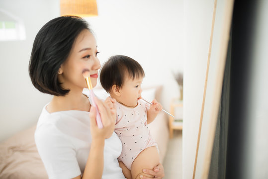 Funny family at home. Mother and her child girl are doing your makeup and having fun near mirror. Baby girl explores mother's cosmetics at home