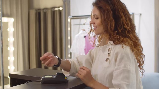 Medium shot of Caucasian young woman paying in clothing store using smart watch, taking shopping bags and going away