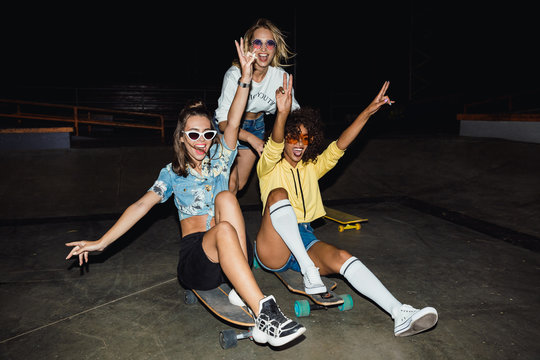 Image of three multinational girls smiling and riding on skateboards at night