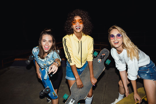 Image of lovely multinational girls holding skateboards at night walk