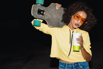 Image of american girl drinking soda and holding skateboard at night